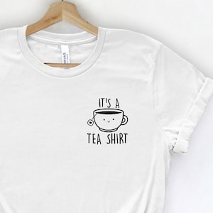 May include: White t-shirt with a black graphic of a teacup with a smiling face and the text "It's a Tea Shirt".
