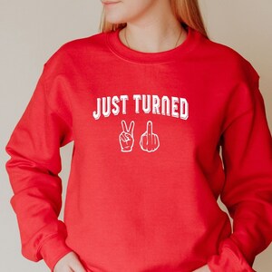 Puede incluir: Sudadera roja con el texto "JUST TURNED" en blanco con un signo de paz y un emoji de dedo medio en blanco.