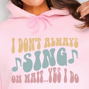 May include: A light pink hoodie with the text "I DON'T ALWAYS SING OH WAIT... YES I DO" in various colors. The design includes musical notes. The hoodie has a drawstring and a front pocket.