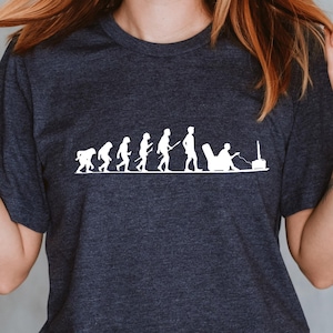 May include: Dark navy blue t-shirt featuring a white graphic depicting the evolution of a person from a primitive form to someone relaxing in a chair. The shirt is paired with blue jeans.
