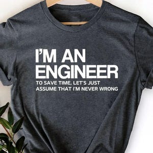 May include: Dark gray t-shirt with the text "I'M AN ENGINEER TO SAVE TIME, LET'S JUST ASSUME THAT I'M NEVER WRONG" in white. The shirt is hanging on a wooden hanger.