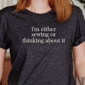 May include: Dark gray t-shirt with rolled sleeves and the text "I'm either sewing or thinking about it" in white. The shirt is paired with blue jeans. A sewing-themed apparel item.