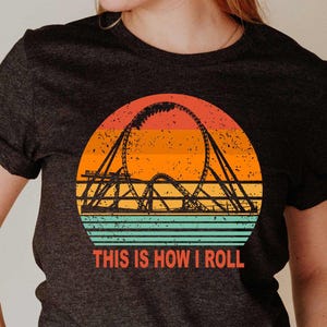 May include: A dark gray t-shirt with a vintage-style graphic of a roller coaster in front of a sunset. The text "This is how I roll" is printed below the graphic.