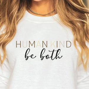 May include: White long-sleeved sweatshirt with the text "HUMAN KIND be both" in a mix of gold and black lettering. The sweatshirt is paired with denim shorts. The overall style is casual and comfortable.