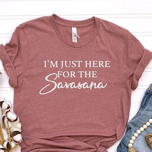 Op de afbeelding: Een mauvekleurig t-shirt met de witte tekst "I'M JUST HERE FOR THE Savasana". Het shirt wordt gecombineerd met een blauwe jeans, een kralenkrans en katoenstelen. De items zijn geplaatst op een wit oppervlak.