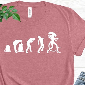 May include: A dusty rose t-shirt with a white graphic depicting the evolution of an alien. The shirt is paired with blue jeans and black sandals. Green leafy plant is in the background.