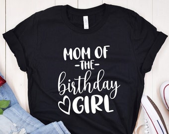May include: Black t-shirt with white text that reads "Mom of the birthday girl" with a heart symbol. The shirt is laid flat on a white wooden surface with a pair of blue jeans and white sneakers.