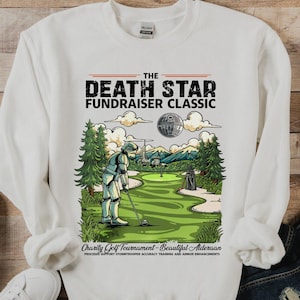 May include: White sweatshirt with a graphic of a Stormtrooper playing golf on a green course, with the Death Star in the background. Text reads "The Death Star Fundraiser Classic". Includes text: "Charity Golf Tournament - Beautiful Alderaan".