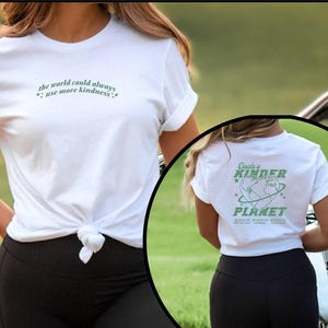 May include: White t-shirt with the text "the world could always use more kindness" in light green. The back of the shirt has the text "Create a Kinder Planet" with a graphic of the Earth. The shirt is tied at the waist.