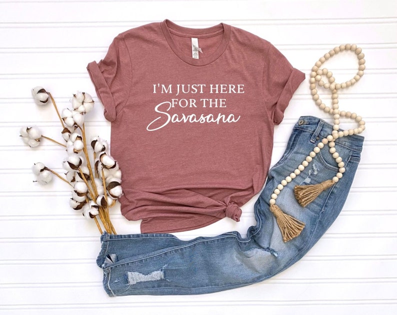 Op de afbeelding: Een mauvekleurig t-shirt met de witte tekst "I'M JUST HERE FOR THE Savasana". Het shirt wordt gecombineerd met een blauwe jeans, een kralenkrans en katoenstelen. De items zijn geplaatst op een wit oppervlak.