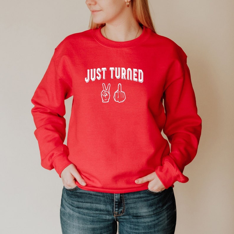 Puede incluir: Sudadera roja con el texto "JUST TURNED" en blanco con un signo de paz y un emoji de dedo medio en blanco.