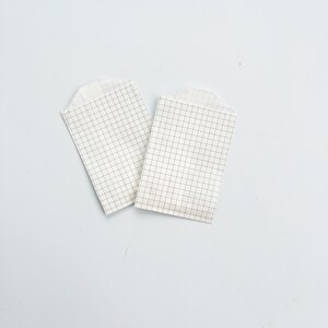 May include: Two white paper bags with a grid pattern. The bags are folded and lying flat on a light blue surface.