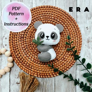 May include: A handmade felt panda bear with gray and white details, holding a green leaf, sits on a woven wicker mat. The image includes text that reads "PDF Pattern + Instructions" in a pink circle.