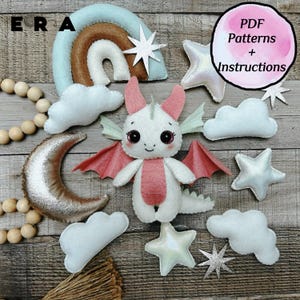 May include: A felt craft kit featuring a white and pink dragon, a rainbow, a silver crescent moon, clouds, and stars. The set includes wooden beads and text that reads PDF Patterns + Instructions, providing the components for a DIY project.