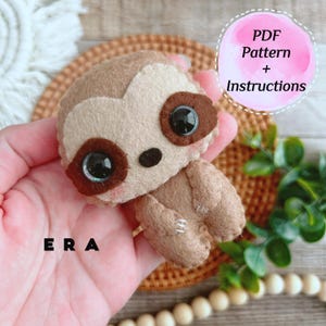 May include: A handcrafted felt sloth toy with large, dark eyes and brown details. The sloth is light brown and tan, with the text "ERA" visible. A pink circle with the text "PDF Pattern + Instructions" is also visible.