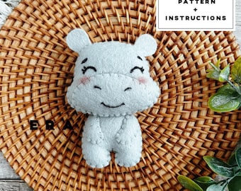 May include: A light grey felt hippo toy with a smiling face and pink cheeks. The hippo has small ears and is stitched together. The image includes text that says "PDF PATTERN + INSTRUCTIONS". The hippo is on a woven brown surface.