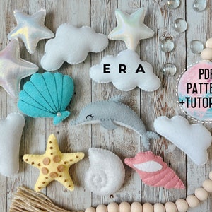 May include: A selection of felt sea-themed decorations, featuring iridescent stars, white clouds, a turquoise seashell, a yellow starfish, a grey dolphin, and pink and white seashells. The word "ERA" is visible, along with a circular graphic that says "PDF PATTERN + TUTORIALS".