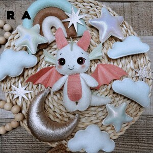 May include: A felt mobile featuring a central white dragon with pink wings and belly, surrounded by light blue clouds, iridescent stars, a silver crescent moon, and a rainbow arch. The text ERA is visible in the upper left corner.