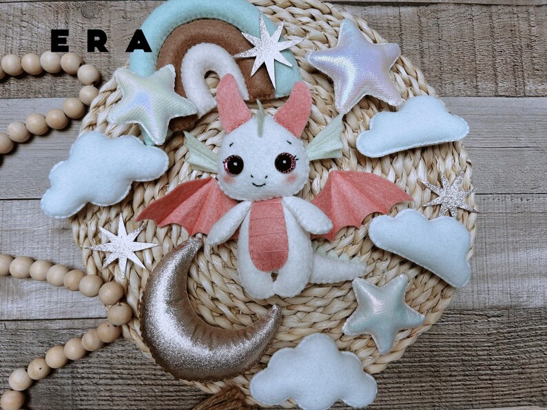 May include: A felt mobile featuring a central white dragon with pink wings and belly, surrounded by light blue clouds, iridescent stars, a silver crescent moon, and a rainbow arch. The text ERA is visible in the upper left corner.
