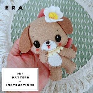 May include: A tan felt dog plushie featuring a white muzzle, black eyes, and a small white flower with a yellow center on its head. The dog has a cream-colored ruffled collar and stitched details. The text ERA and PDF PATTERN + INSTRUCTIONS are visible.