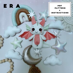 May include: A felt craft kit featuring a white dragon with coral wings, a rainbow, clouds, stars, and a crescent moon. The text reads ERA PDF PATTERN + INSTRUCTIONS. The set includes various soft shapes in pastel colours, including a silver moon and stars.