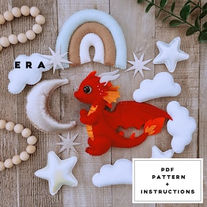 Puede incluir: Un kit de móvil de fieltro con un dragón rojo, un arcoíris, estrellas, nubes y una luna. El kit incluye un patrón PDF e instrucciones. La palabra "ERA" también es visible.