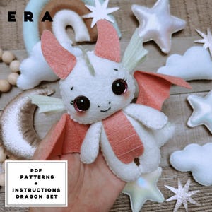 May include: A white felt dragon plush toy with coral-coloured wings, horns, and belly accents, featuring large dark eyes with pink highlights. The text "ERA" appears in the top left, and a box reads "PDF PATTERNS + INSTRUCTIONS DRAGON SET".