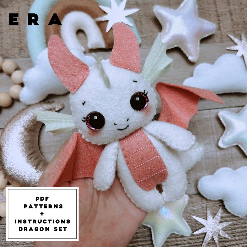 May include: A white felt dragon plush toy with coral-coloured wings, horns, and belly accents, featuring large dark eyes with pink highlights. The text "ERA" appears in the top left, and a box reads "PDF PATTERNS + INSTRUCTIONS DRAGON SET".