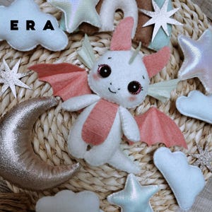 May include: A white and pink felt dragon plush toy surrounded by felt clouds, stars, and a crescent moon. The dragon has large dark eyes with pink accents, small wings, and a pink belly. The text ERA is visible in the upper left corner.
