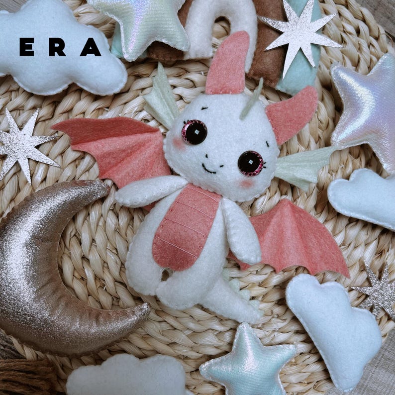 May include: A white and pink felt dragon plush toy surrounded by felt clouds, stars, and a crescent moon. The dragon has large dark eyes with pink accents, small wings, and a pink belly. The text ERA is visible in the upper left corner.