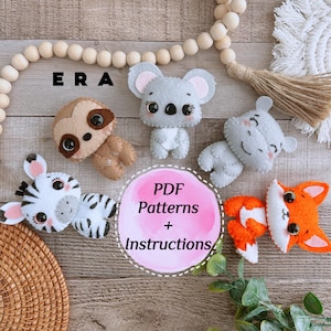 Op de afbeelding: Een verzameling handgemaakte vilten dierenornamenten, waaronder een koala, een luiaard, een zebra, een vos en een nijlpaard. De ornamenten zijn in verschillende kleuren, met de koala en het nijlpaard in grijs, de vos in oranje en wit en de zebra in wit en zwart. De tekst "PDF Patterns + Instructions" is inbegrepen.