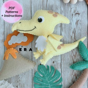 May include: A yellow felt dinosaur with a white wing and brown spots. The dinosaur is sitting on a wooden surface with a green felt leaf, a brown felt volcano, and a gray felt cloud.