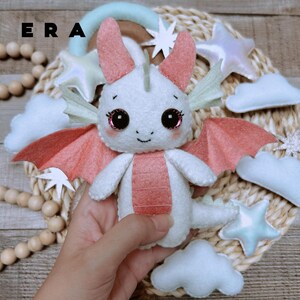 May include: A white and coral felt dragon plush toy with large black eyes, pointed ears, and wings. The dragon features a coral-coloured belly and a small tail. The text ERA is visible in the upper left corner of the image, which includes decorative felt stars and clouds.