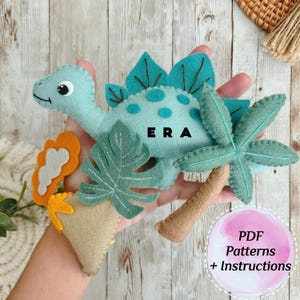 May include: A felt dinosaur toy with green and blue felt, a green felt palm tree, a green felt monstera leaf, and a tan felt volcano. The dinosaur has the name "ERA" embroidered on its back. The text "PDF Patterns + Instructions" is on a pink background.