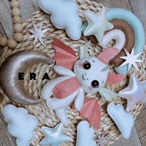 May include: A felt mobile featuring a white and pink dragon with large dark eyes, surrounded by white clouds, iridescent stars, a brown and mint green rainbow, and a textured gold crescent moon. The letters ERA are visible in black.