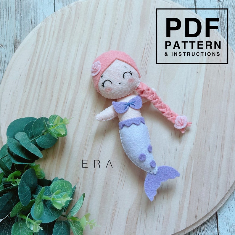 May include: A felt mermaid doll with pink hair, a purple tail, and a purple bow. The doll is smiling and has a pink flower on her head. The doll is on a wooden background with the text "PDF PATTERN & INSTRUCTIONS" and "ERA" visible.