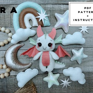 May include: A felt dragon plush toy surrounded by felt clouds, stars, a crescent moon, and a rainbow. The dragon is white with pink wings and belly. A string of wooden beads is on the left. Text in the image reads: ERA, PDF PATTERNS + INSTRUCTIONS.