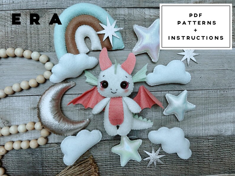 May include: A felt dragon plush toy surrounded by felt clouds, stars, a crescent moon, and a rainbow. The dragon is white with pink wings and belly. A string of wooden beads is on the left. Text in the image reads: ERA, PDF PATTERNS + INSTRUCTIONS.