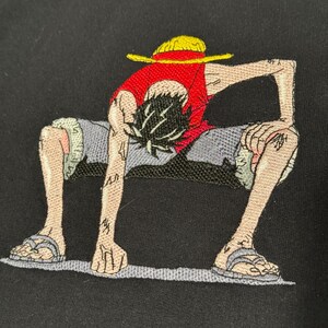 May include: A black t-shirt featuring an embroidered graphic of a character with black hair wearing a yellow straw hat, a red sleeveless shirt, and grey shorts. The character is in a crouching position with hands touching the ground.