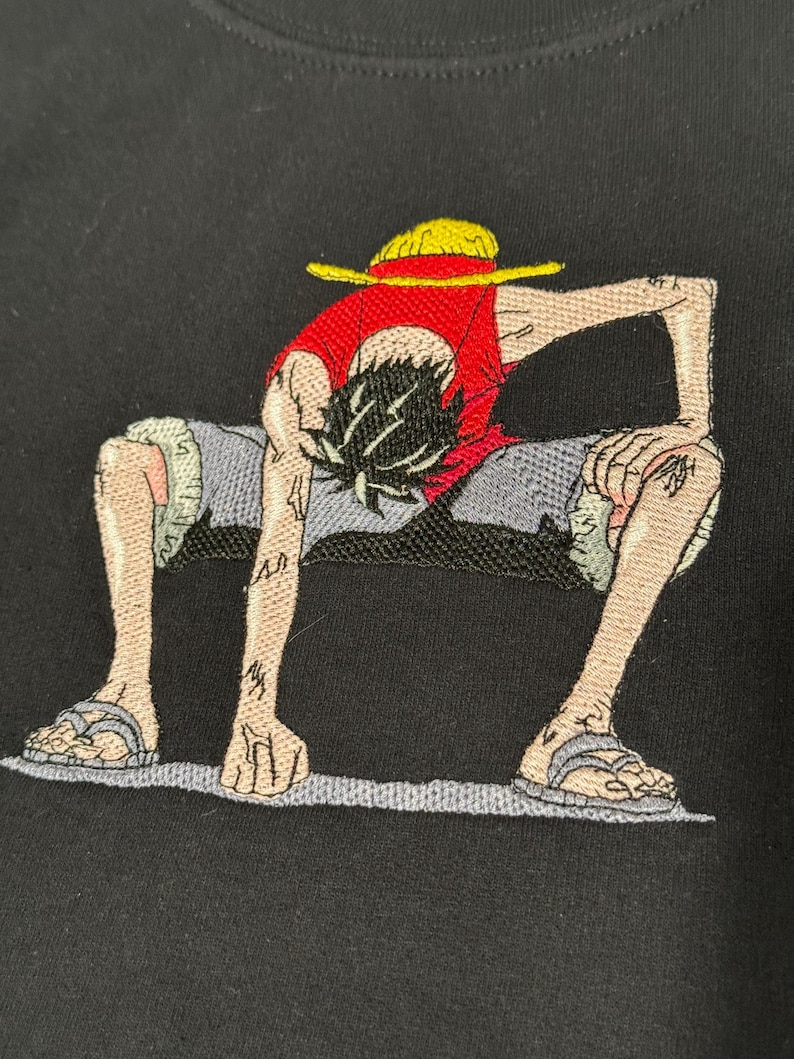 May include: A black t-shirt featuring an embroidered graphic of a character with black hair wearing a yellow straw hat, a red sleeveless shirt, and grey shorts. The character is in a crouching position with hands touching the ground.