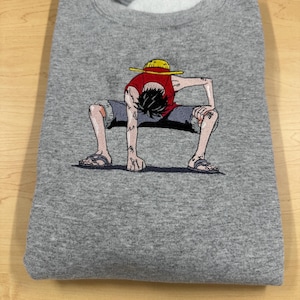 May include: A heather gray crewneck sweatshirt featuring a detailed embroidered graphic of a cartoon character wearing a yellow straw hat, a red sleeveless shirt, and gray shorts, crouched in a low stance with hands touching the ground.