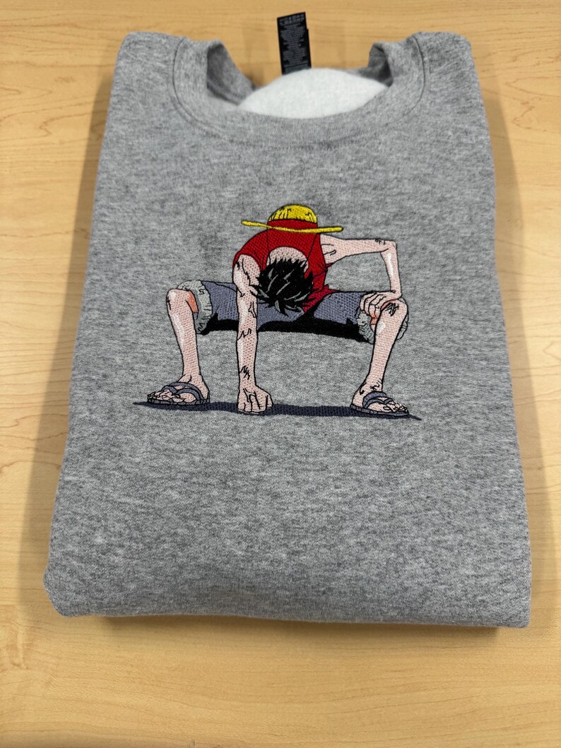 May include: A heather gray crewneck sweatshirt featuring a detailed embroidered graphic of a cartoon character wearing a yellow straw hat, a red sleeveless shirt, and gray shorts, crouched in a low stance with hands touching the ground.