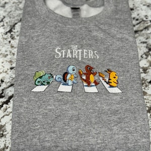 Könnte beinhalten: Grauer Pullover mit einem weißen gestickten Design der Pokémon-Charaktere Bisasam, Schiggy, Glumanda und Pikachu, die über einen Zebrastreifen laufen, mit dem Text "The Starters" darüber.