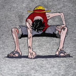 May include: A heather gray sweatshirt featuring a detailed embroidered graphic of a cartoon character with black hair, wearing a yellow straw hat, a red sleeveless shirt, and blue shorts, crouched in a low stance with one hand on the ground.