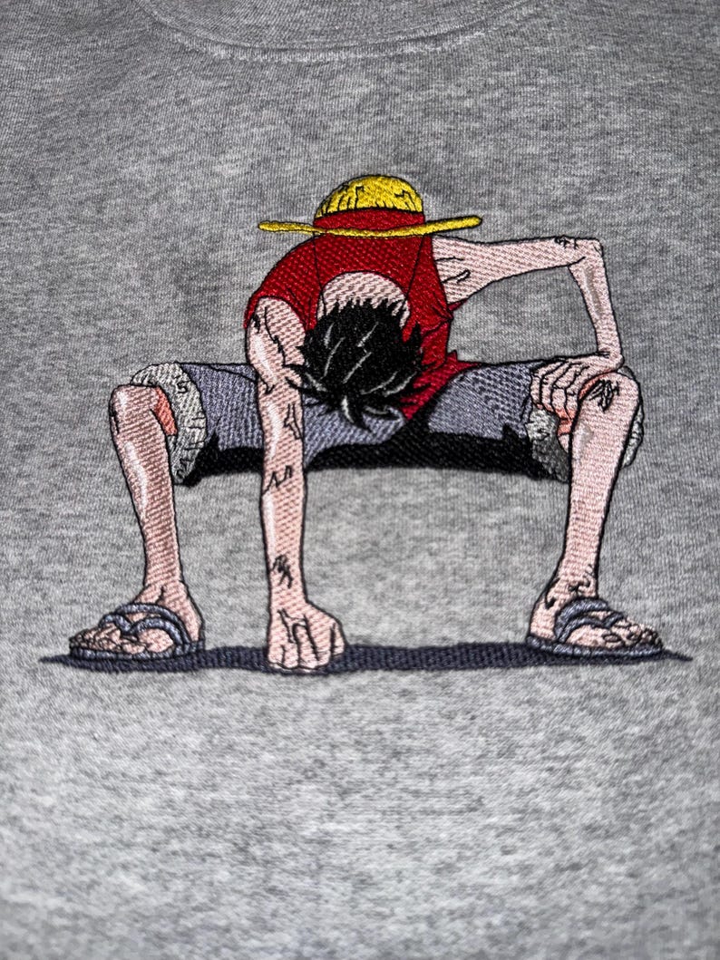 May include: A heather gray sweatshirt featuring a detailed embroidered graphic of a cartoon character with black hair, wearing a yellow straw hat, a red sleeveless shirt, and blue shorts, crouched in a low stance with one hand on the ground.
