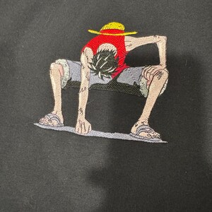 May include: A black sweatshirt featuring an embroidered graphic of a character in a crouching pose. The character wears a red vest, blue shorts, and a yellow straw hat. The figure has black hair, tan skin, and is wearing sandals on their feet.