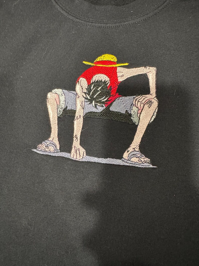 May include: A black sweatshirt featuring an embroidered graphic of a character in a crouching pose. The character wears a red vest, blue shorts, and a yellow straw hat. The figure has black hair, tan skin, and is wearing sandals on their feet.