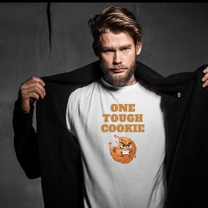 May include: A white t-shirt with the text "ONE TOUGH COOKIE" in brown letters and a cartoon cookie with an angry face.