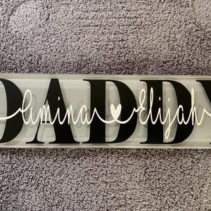 May include: A rectangular sign with the word "DADDY" in large black block letters. The names "Elimina" and "Elijah" are written in white cursive script, with a heart symbol between them.