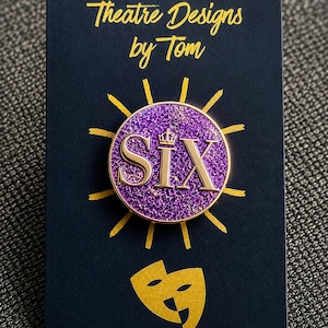 Op de afbeelding: Een ronde paarse glitter emaille speld met de gouden tekst SIX en een klein kroontje boven de letter I. De speld is bevestigd op een zwarte kaart met de gouden tekst Theatre Designs by Tom en een icoon van een theatermasker.